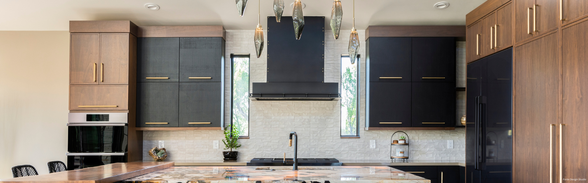 Modern kitchen featuring Banner Leadville Custom dark wood cabinets with gold handles, a black range hood, built-in ovens, marble island countertop, pendant lights, and tall windows for natural light.