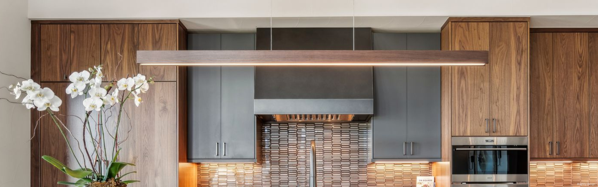 Banner Mesa Custom Modern kitchen featuring Banner Mesa Custom wood cabinets, a tiled backsplash, stainless steel oven, and a large rectangular pendant light above the sink. A white orchid decorates the left side of the counter.