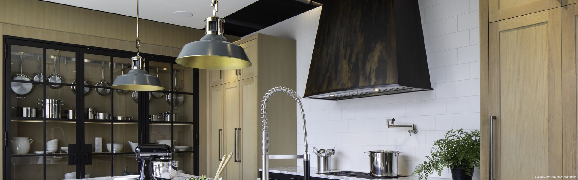 Modern kitchen featuring Banner Tabernash Custom light wood cabinets, a black range hood, industrial pendant lights, a glass-fronted cabinet with displayed cookware, and a sleek faucet near a green potted plant on the counter.