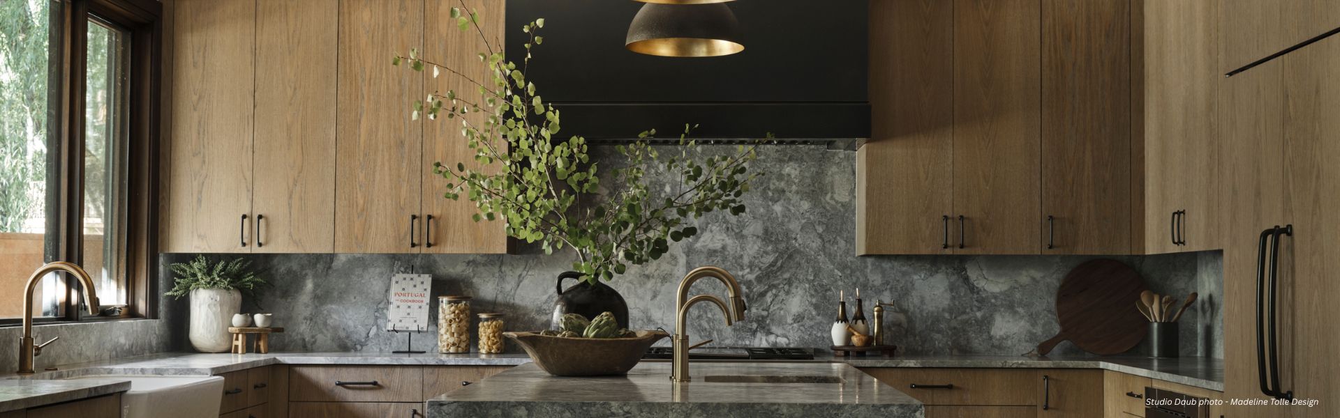 EVERGREEN Box Tailored √ Modern kitchen with light wood cabinets, gray stone counters, gold hardware, and two gold pendant lights. An EVERGREEN Box Tailored √ filled with green branches sits on the island, adding a natural touch.