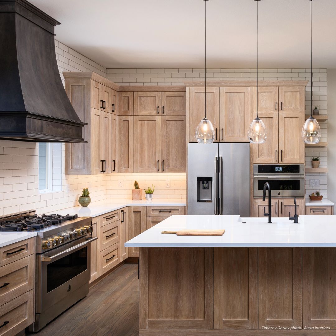 The Montrose-essential-gallery1√ features a spacious, modern kitchen with light wood cabinets, white countertops, stainless steel appliances, a large island, three glass pendant lights, white subway tile backsplash, and a contrasting dark range hood.