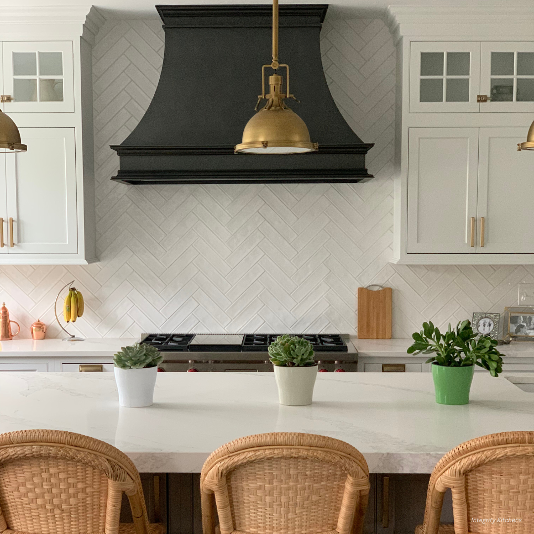The Montrose-essential-gallery2√ features a modern kitchen with a white herringbone backsplash, black range hood, gold pendants, white cabinets, marble island with potted plants, and woven bar stools.
