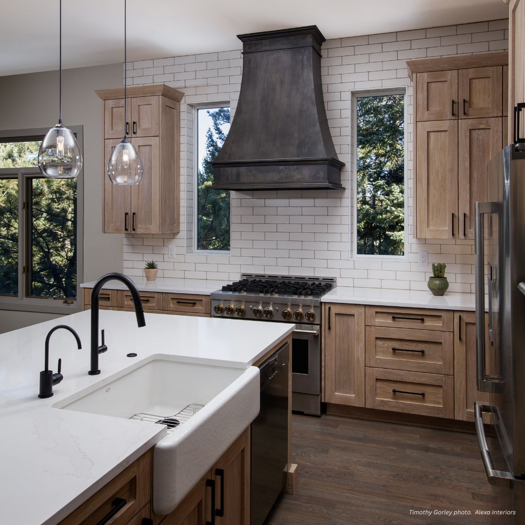 Modern kitchen featuring Montrose-essential-gallery3: light wood cabinets, white countertops, farmhouse sink, black fixtures, stainless steel stove with vent hood, subway tile backsplash, and windows with a view of trees.