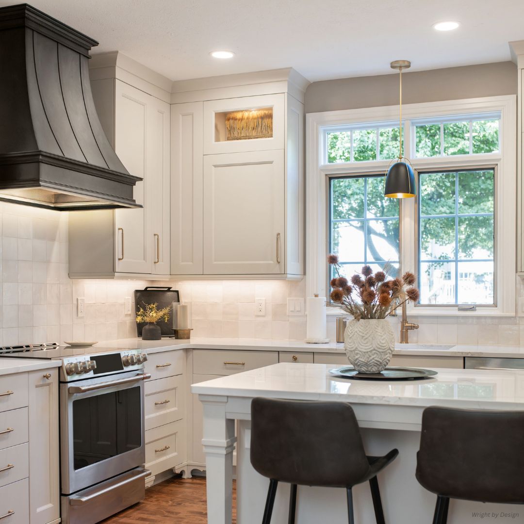 The Montrose-refined-gallery2√ features a modern kitchen with white cabinets, stainless steel oven, large black range hood, island seating for two, pendant lighting, a vase of dried flowers, and a large window for natural light.