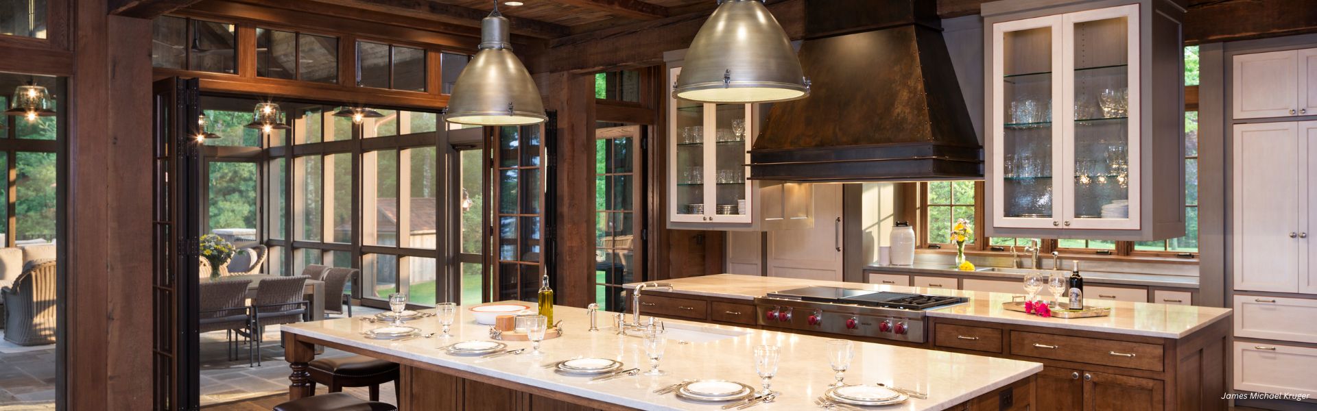 RU_Banner_Content-4 The RU_Banner_Content-4 features a spacious modern kitchen with a large island, wood and glass-front cabinets, pendant lighting, expansive windows overlooking greenery, and a dining table set with plates and glasses.