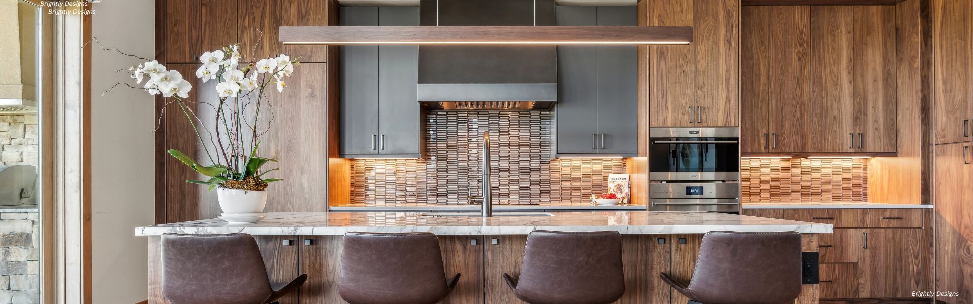 RU_Banner_Content-5 RU_Banner_Content-5 features a modern kitchen with wood cabinets, a marble island and four brown chairs, an orchid plant, built-in appliances, tile backsplash, and bright under-cabinet plus pendant lighting.