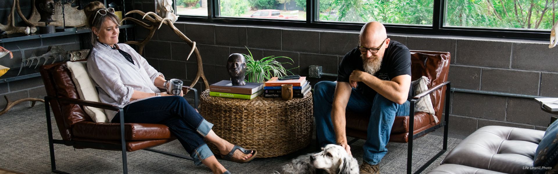 In a cozy, modern lounge with leather chairs and a woven table, a man and woman relax as natural light streams in through large windows. The RU_Banner_Content-7 decorates the space alongside books, a plant, and a bust on the table.