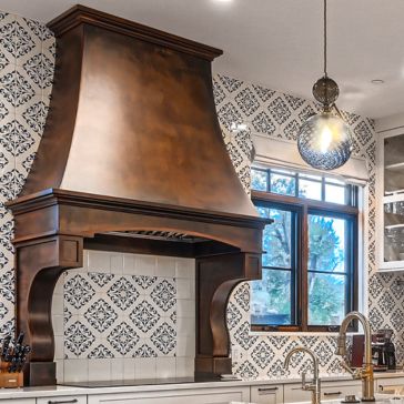 Rustic Iron-2 A kitchen features a large Rustic Iron-2 range hood above the stove, set against patterned tile walls. A window, modern light fixture, and part of a sink and countertop are also visible in the bright, stylish space.