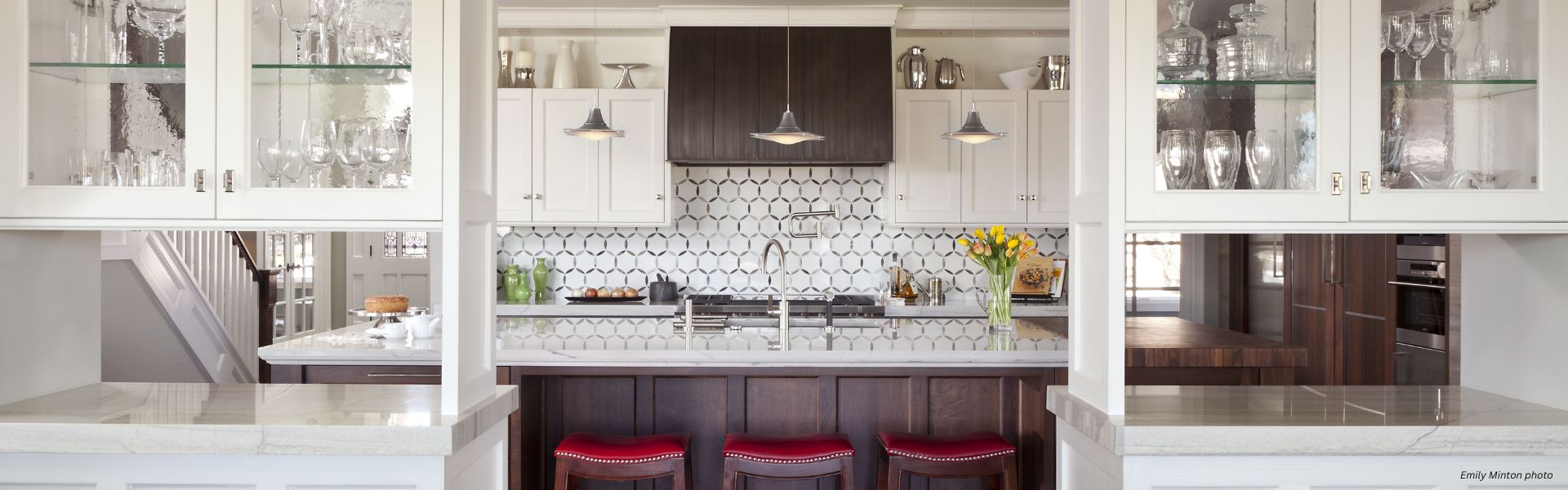 The Snowmass-3 features a modern kitchen with white cabinets, glass shelves for glassware, a dark wood island with three red cushioned stools, stainless steel appliances, patterned tile backsplash, and vases of yellow flowers on the counter.