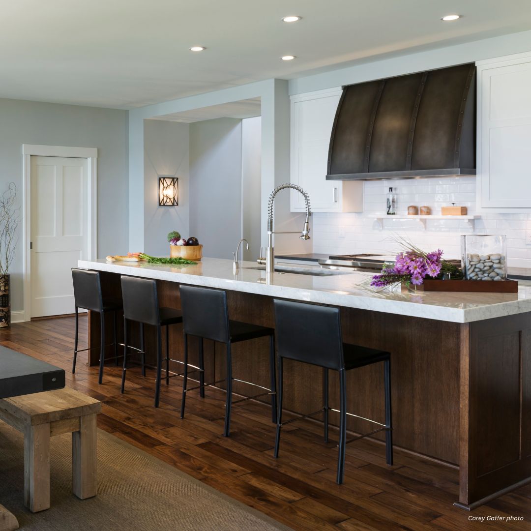 aspen-refined-gallery3 √ The aspen-refined-gallery3 showcases a modern kitchen with a large island, white countertop, wooden base, three black bar stools, gooseneck faucet, flowers, baskets on the island, wood flooring, and recessed lighting.