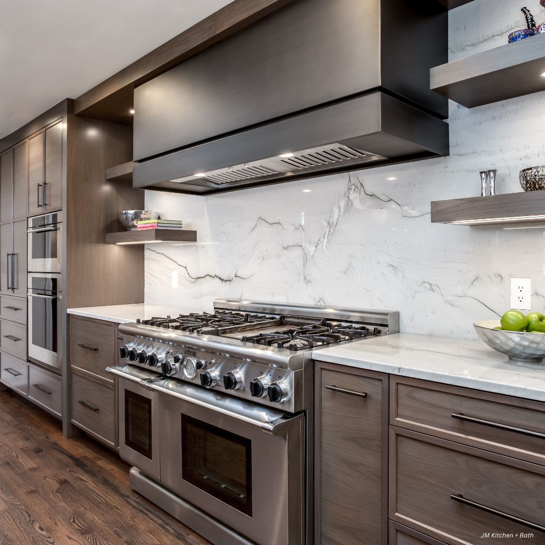 box-tailored-gallery1√ Box-tailored-gallery1√ features a modern kitchen with a stainless steel stove and oven, marble backsplash, wood cabinetry, open shelves adorned with decor, and a bowl of green apples on the white countertop.