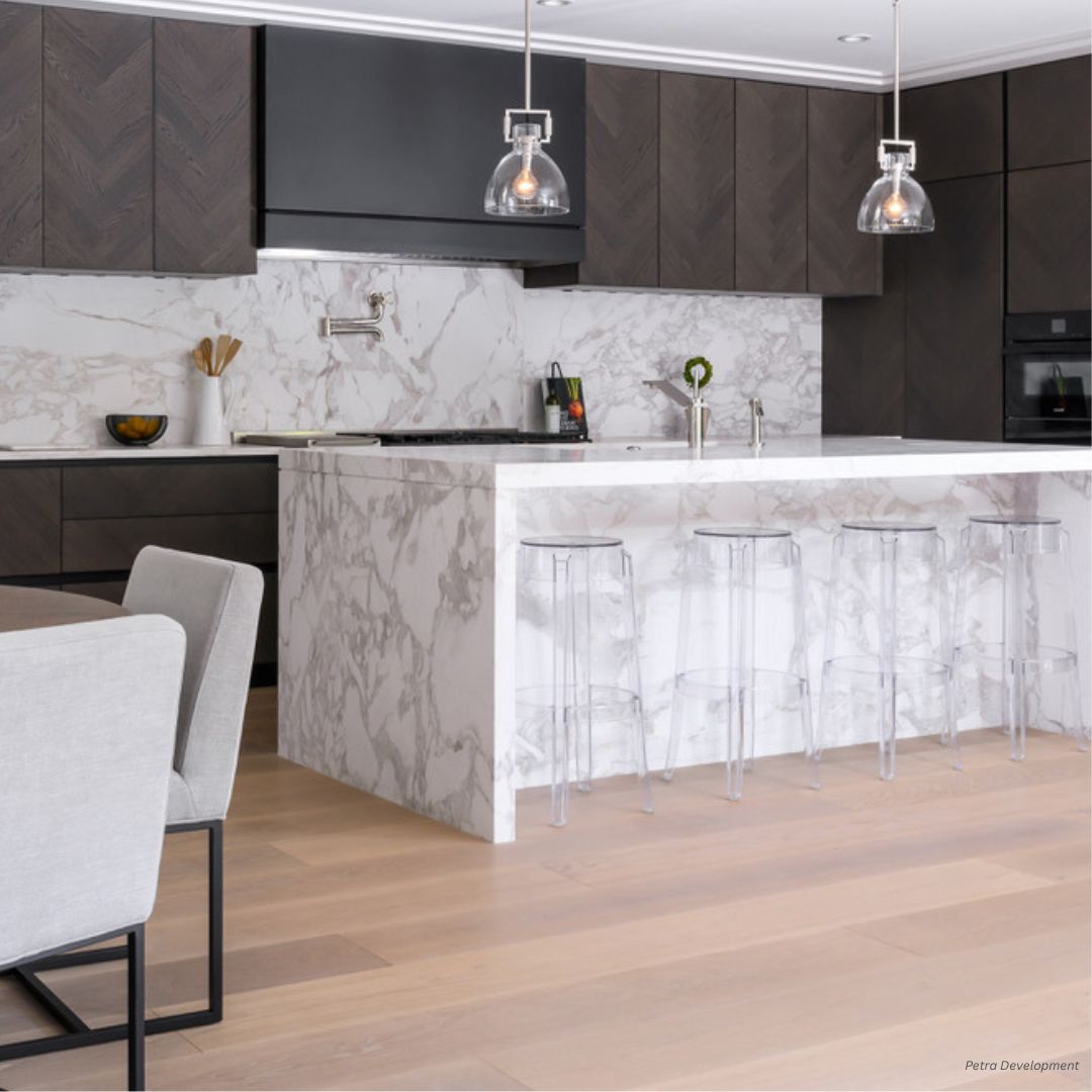 box-tailored-gallery2√ The modern kitchen features box-tailored-gallery2√ dark wood cabinets, a white marble backsplash and island, clear acrylic barstools, pendant lights above the island, light wood flooring, and a dining table with light upholstered chairs in the foreground.