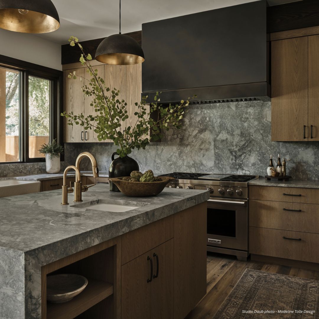 box-tailored-gallery3√ The box-tailored-gallery3√ showcases a modern kitchen with gray stone counters and backsplash, wood cabinets, a stainless steel stove, black range hood, brass fixtures, pendant lights, and a vase with green branches on the island lit by sunlight.