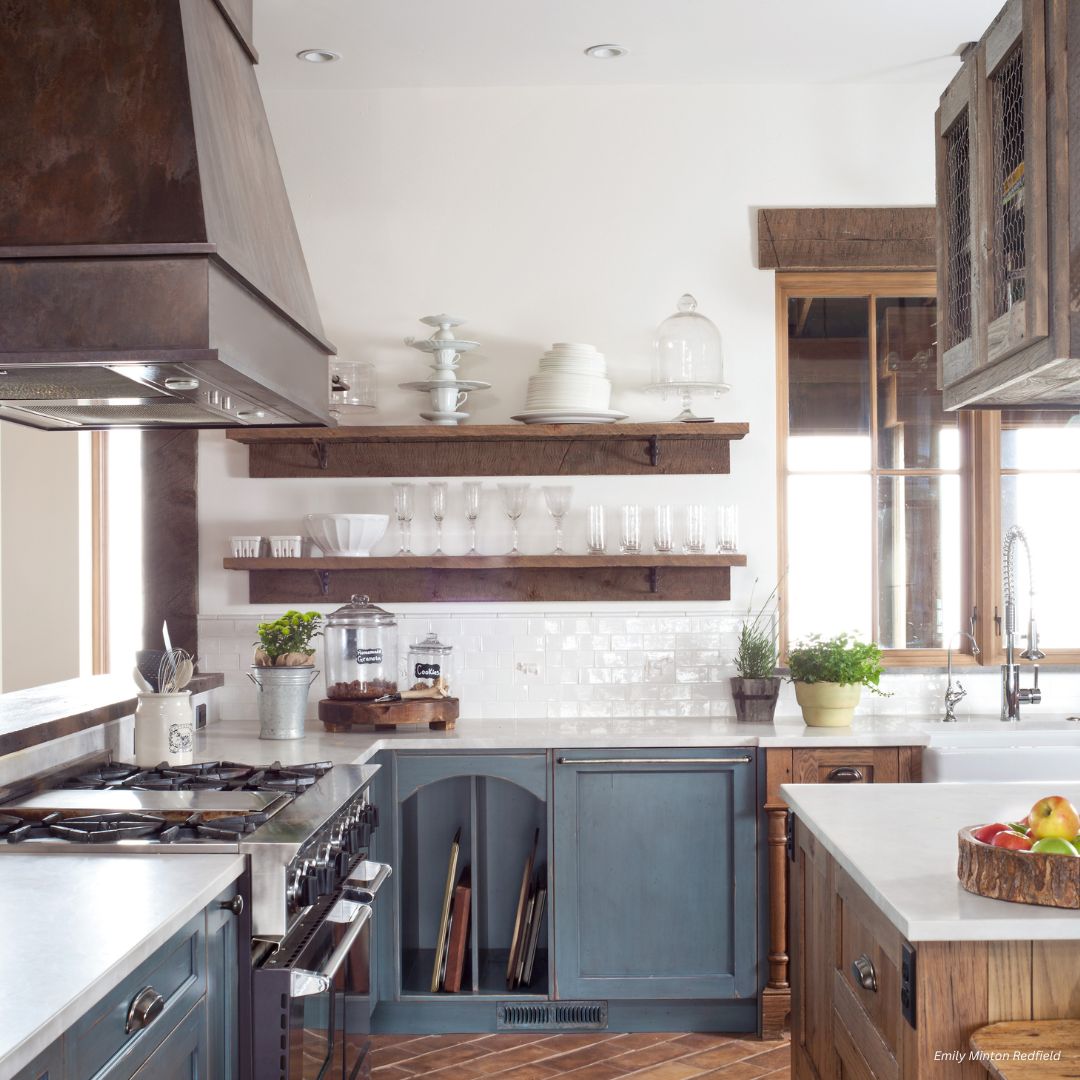 creed-essential-gallery2√ The creed-essential-gallery2√ showcases a bright kitchen with blue cabinets, open wooden shelves displaying dishes, a stainless steel stove, potted herbs on the counters, apples in a bowl, and sunlight pouring through a large window.