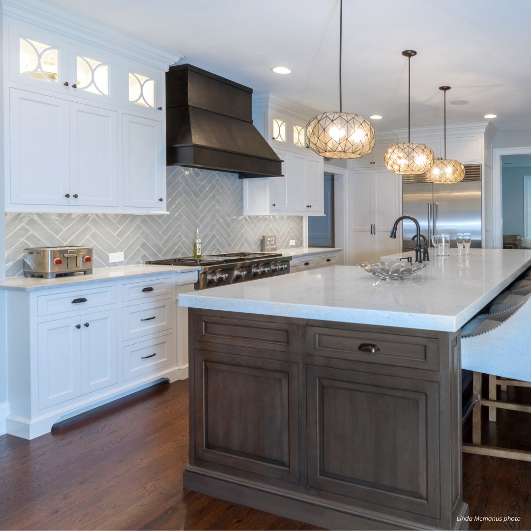 creed-tailored-gallery1√ The creed-tailored-gallery1√ features a modern kitchen with white cabinets, a spacious gray island, stainless steel appliances, dark range hood, herringbone tile backsplash, pendant lights, wooden floors, and island seating.
