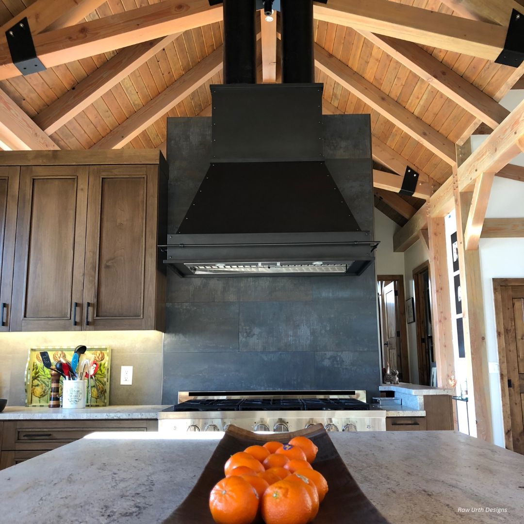 creede-classic-gallery2√ A modern kitchen featuring the creede-classic-gallery2√ with a vaulted wood ceiling, black range hood, wooden cabinets, and a tray of oranges on the stone island countertop in the foreground.