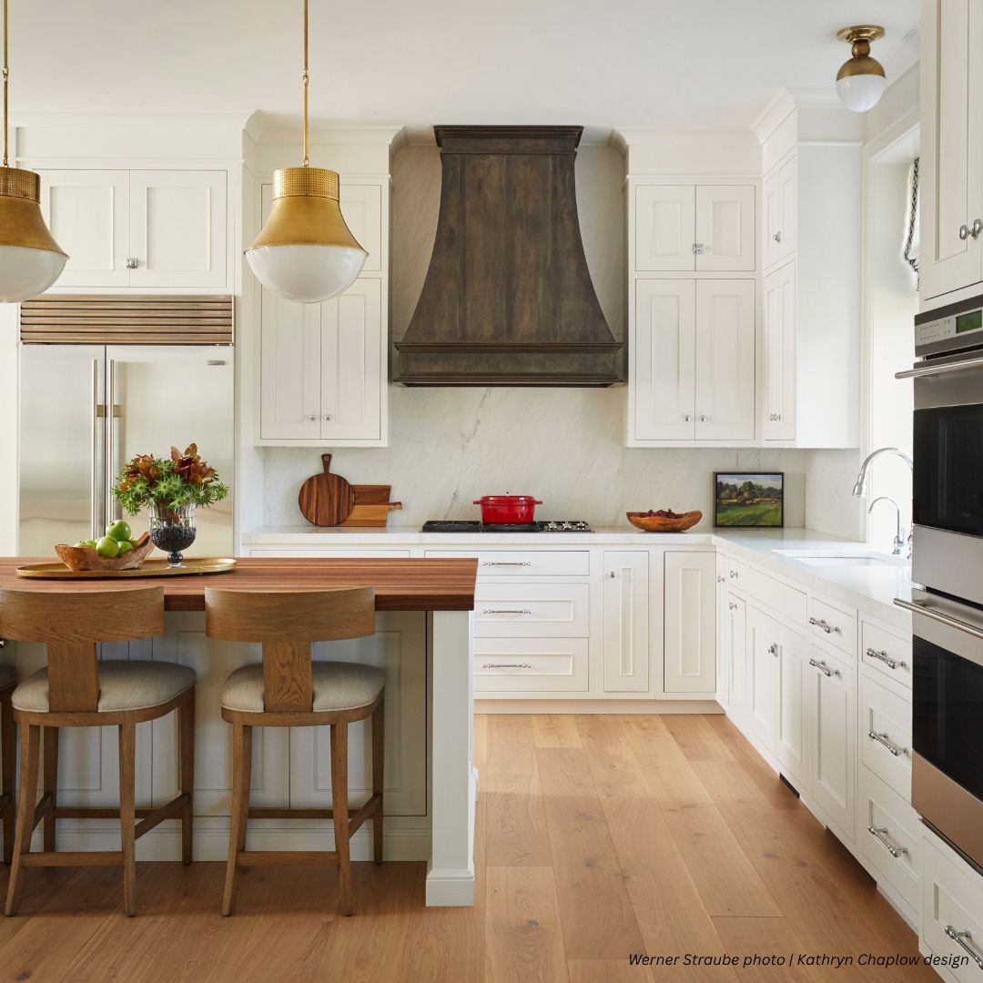 montrose-classic-gallery 1√ A bright, modern kitchen featuring the montrose-classic-gallery 1√: white cabinets, dark wood range hood, stainless steel appliances, wood floors, built-in oven, and a kitchen island with two stools and a wooden countertop.
