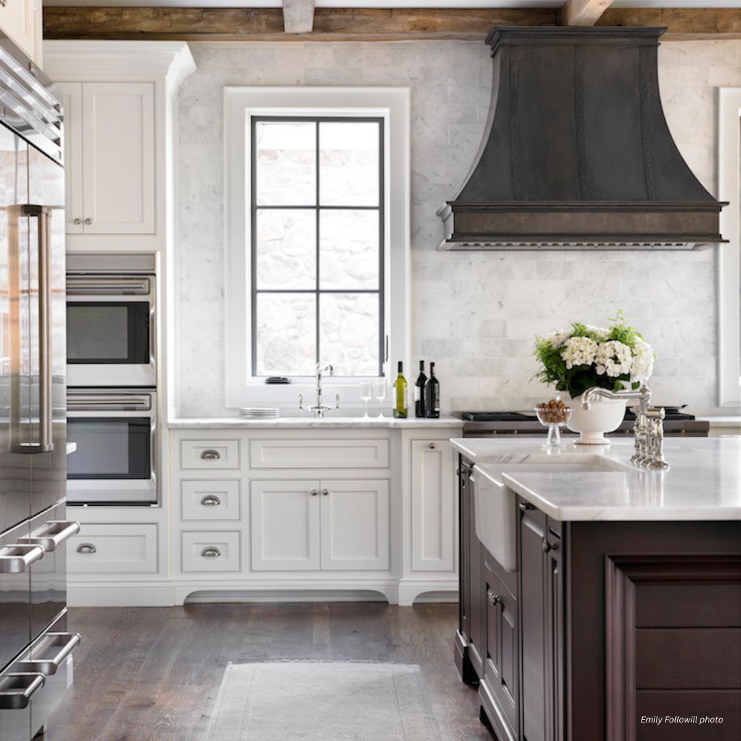 montrose-classic-gallery2√ The montrose-classic-gallery2√ kitchen features white cabinetry, a dark island, stainless steel appliances, rustic beams, a marble backsplash, and a large window accented by fresh flowers and bottles on the countertop.