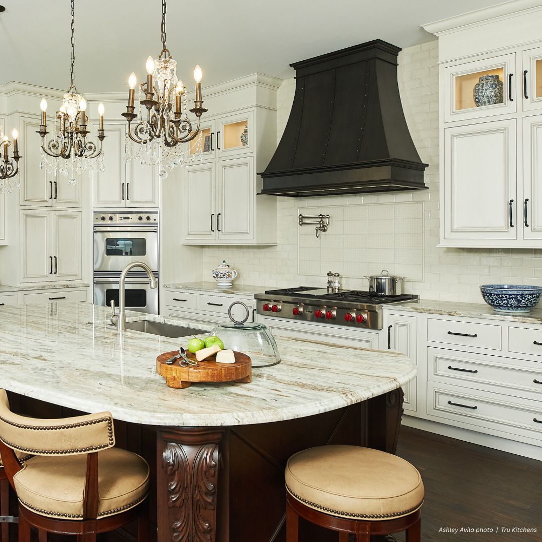 The Montrose Refined Gallery1√ kitchen features white cabinets, a marble island countertop with a dark wood base, beige cushioned stools, stainless steel appliances, a black range hood, chandeliers, and blue-and-white pottery accents.