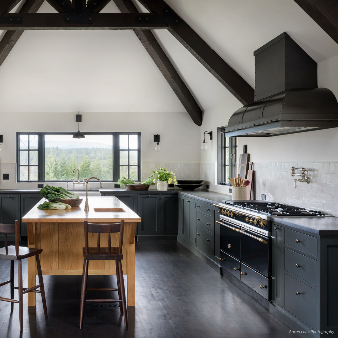 palisade gallery2 The palisade gallery2 features a modern farmhouse kitchen with black cabinets, a wooden island with two chairs, exposed dark ceiling beams, large windows, a black range and hood, plus greenery on the counter.