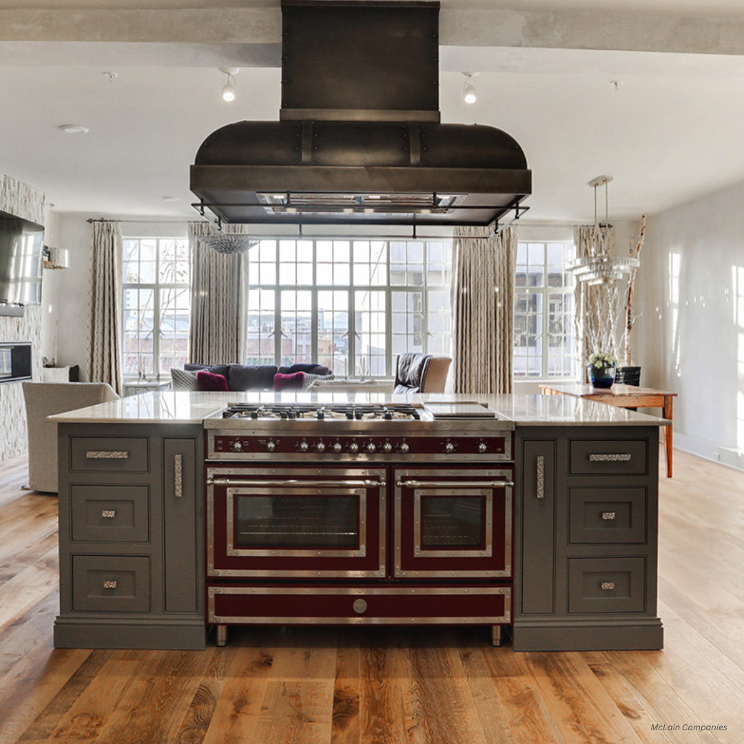 palisade gallery3 A modern kitchen features a central island with a palisade gallery3 dark red and stainless steel stove, grey cabinetry, wood floors, large windows, neutral walls, a black vent hood above, and the living area in the background.