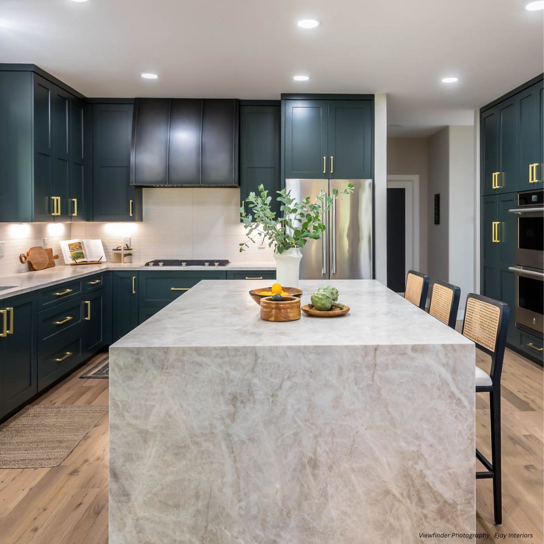 snowmass-classic-gallery 2√ Modern kitchen featuring snowmass-classic-gallery 2√ cabinets in dark teal, gold hardware, marble island with greenery, built-in oven, stainless steel fridge, light wood floors, and three island chairs.