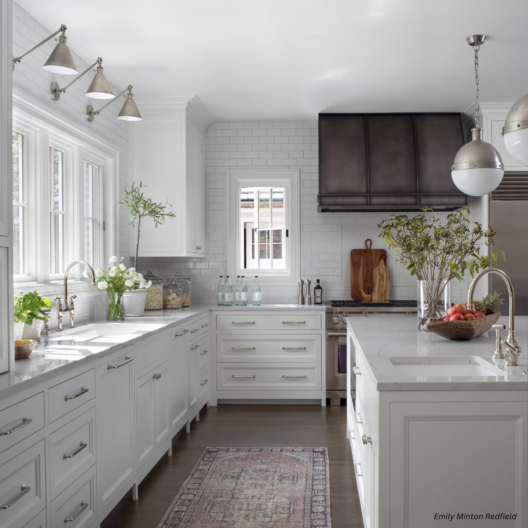 snowmass-refined-gallery1√ The snowmass-refined-gallery1√ features a bright, modern kitchen with white cabinets, marble countertops, stainless steel appliances, a dark range hood, large windows, and neatly arranged fresh flowers, fruit, and essentials on the counters.