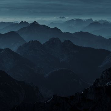82 captures a succession of dark, misty mountain ranges beneath a cloudy, moody sky, with layers of blue and gray peaks fading into the distance.