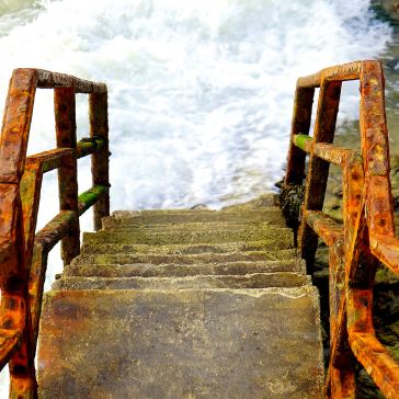 84 The 84 features stone steps with rusted railings descending into foamy, churning water, evoking a river or sea access; moss covers the lower rails.