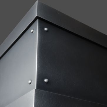 Close-up of the corner of an Antique 6 black metal box with rivets, set against a plain gray background. The image highlights its sharp edges and smooth, matte metal surface.