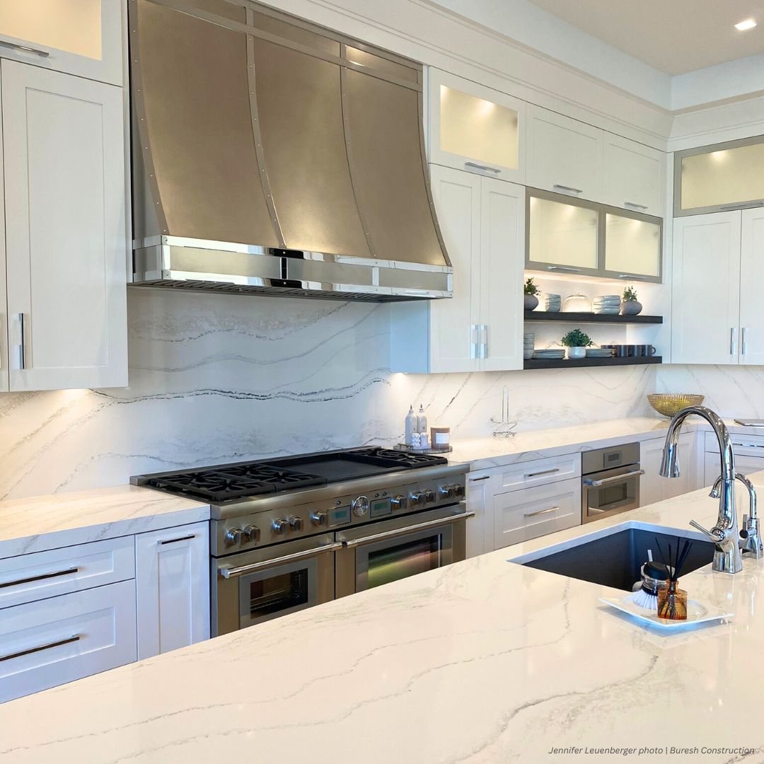 Avon gallery1 ✅ The Avon gallery1 features a modern kitchen with white cabinets, stainless steel range and hood, marble countertops and backsplash, built-in oven, open shelves with decor, and a spacious island with sink and curved faucet.