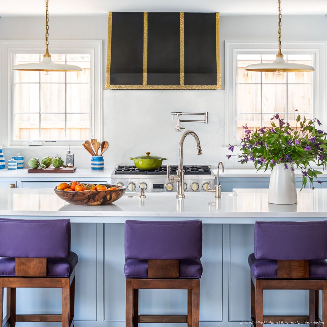 Avon gallery2 The Avon gallery2 features a bright kitchen with a large island, three purple-cushioned stools, white countertop, fruit bowl, flower vase, modern appliances, black and gold range hood, and two pendant lights.