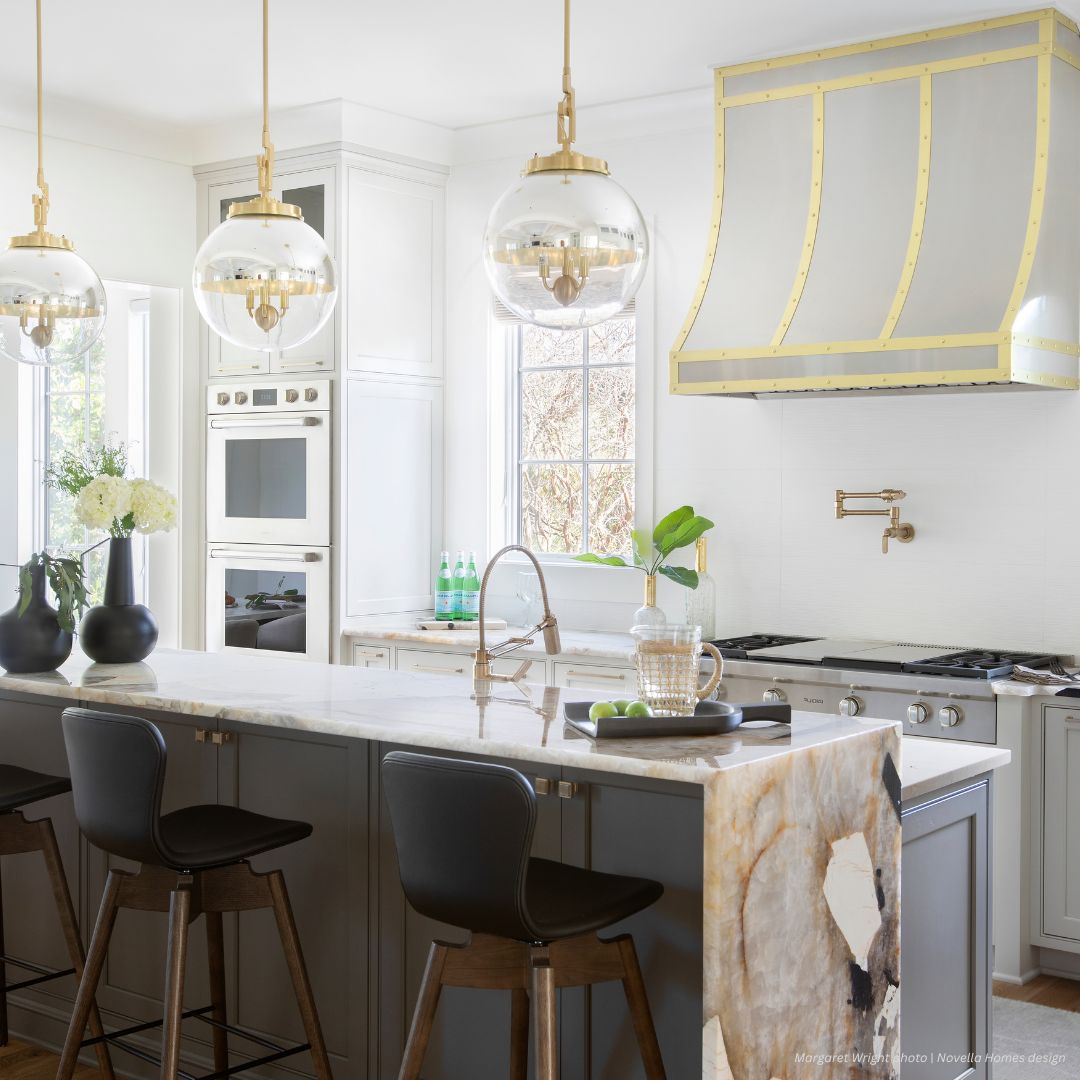 Avon gallery3 The Avon gallery3 kitchen features a contemporary design with a marble island, three black barstools, globe pendant lights, gold-trimmed range hood, built-in ovens, and expansive windows for abundant natural light.