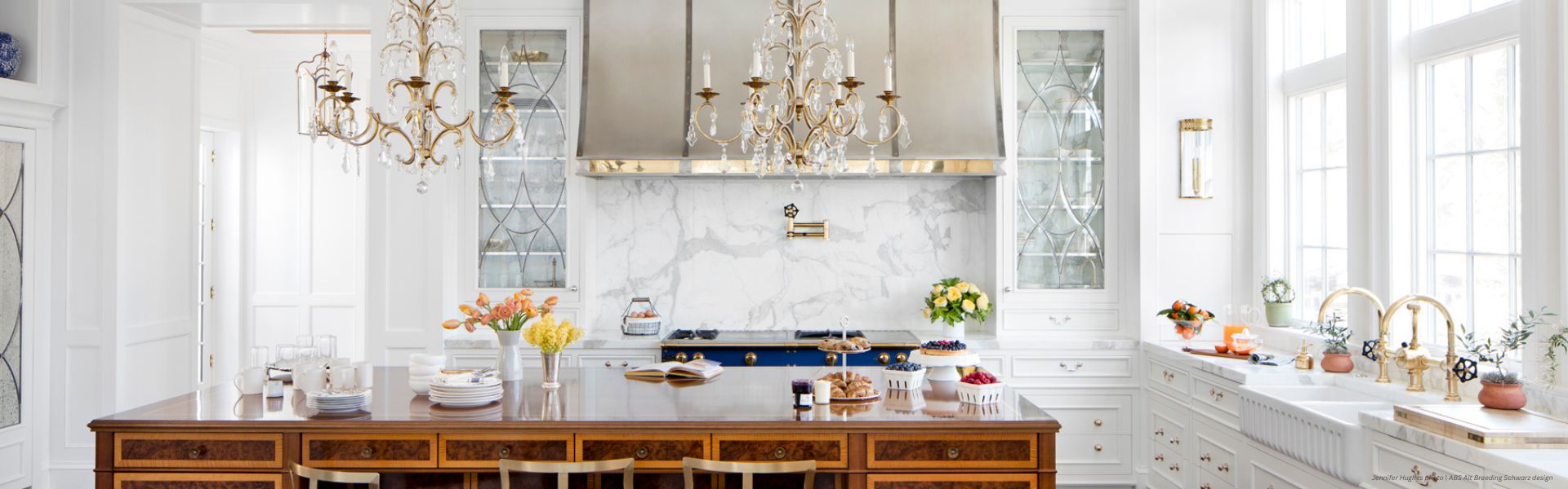 Banner Avon Custom Bright, elegant kitchen featuring the Banner Avon Custom island, gold chandeliers, white cabinets, marble backsplash, and brass fixtures. Cakes, flowers, and fruit decorate the counters as sunlight pours through tall windows.