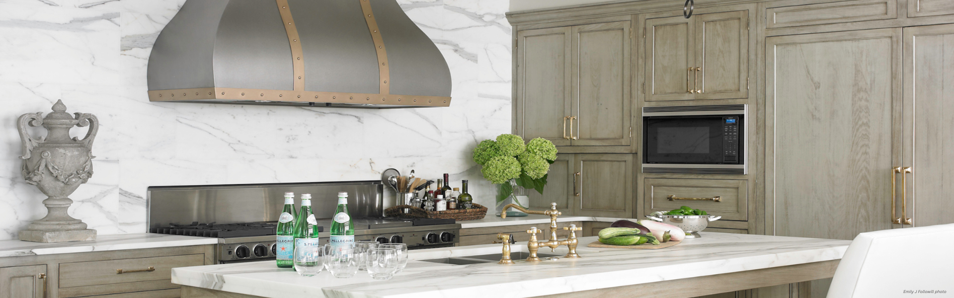 Banner Bellvue Custom ✅ A modern kitchen featuring Banner Bellvue Custom marble countertops and backsplash, light wood cabinets, a large range hood, built-in microwave, green hydrangeas, bottled water, glassware, and fresh vegetables on the island.