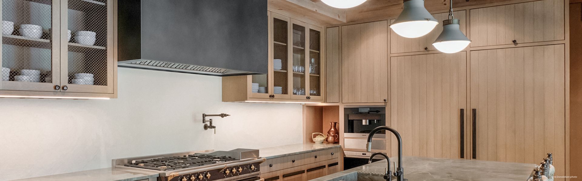 Banner Box Custom ✅ Modern kitchen featuring Banner Box Custom cabinets in light wood, stainless steel stove, glass-fronted doors with dishes, wall-mounted pot filler, built-in appliances, and pendant lights over a marble island with sink.