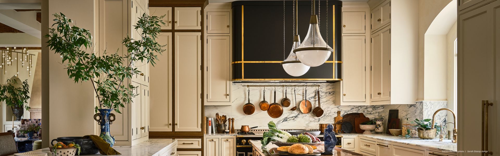 The Banner Broadmoor Custom kitchen features cream cabinets, marble countertops, globe pendants over a central island with bread and vegetables, stylish copper pots, green plants, and large windows for natural light.