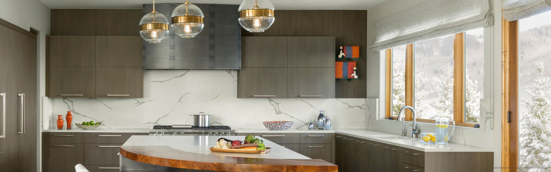Banner Gunnison Custom Modern kitchen featuring Banner Gunnison Custom dark wood cabinets, white marble backsplash and countertops, large windows with snowy views, plus a kitchen island with wooden edge and pendant lighting overhead.