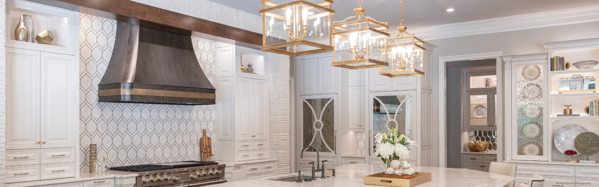 Banner Mary Jane Custom ✅ A stylish kitchen features Banner Mary Jane Custom cabinets, a large marble island, stainless steel range hood, gold pendant lights, decorative shelves with vases and plates, and fresh flowers on the countertop.