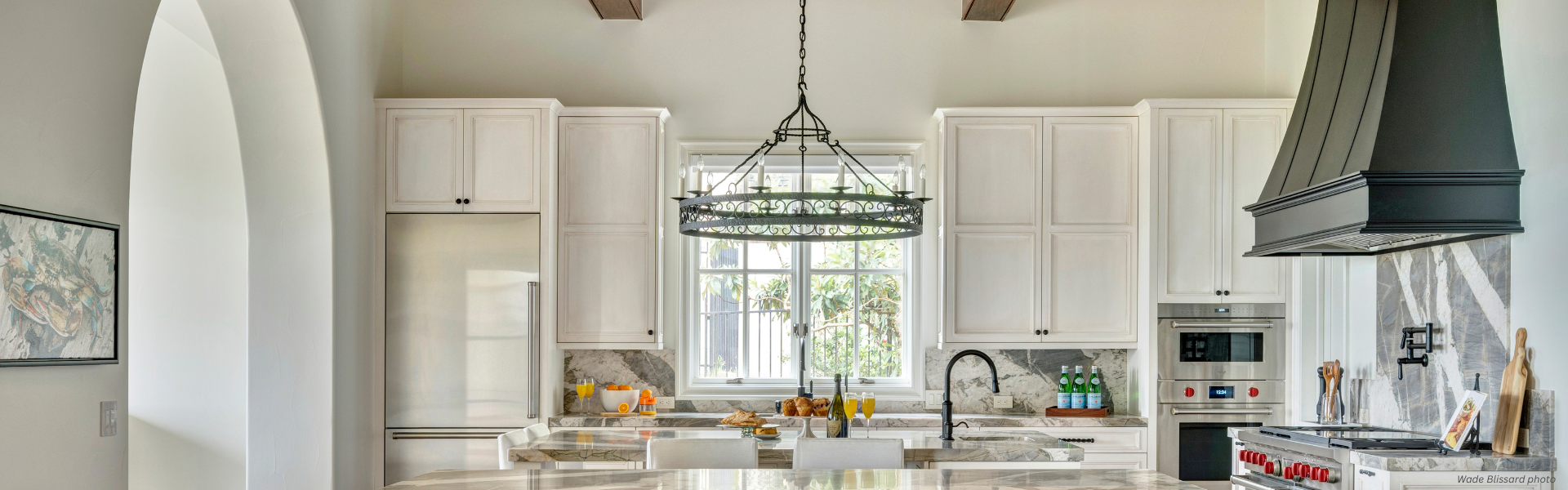 Banner Montrose Custom ✅ Bright, modern kitchen featuring Banner Montrose Custom white cabinets, marble countertops, a central island with stools, large chandelier, stainless steel appliances, and a window above the sink for natural light.