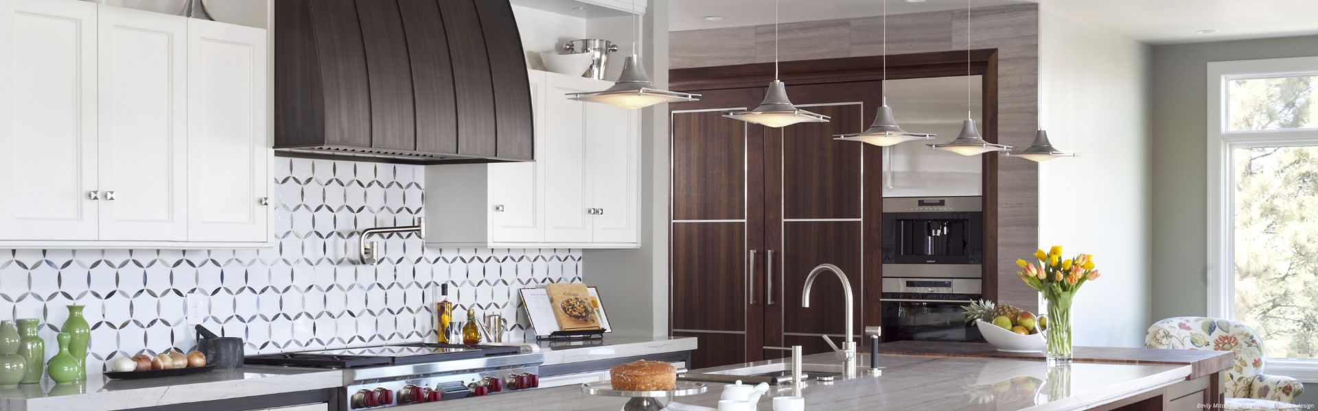Banner Snowmass Custom Modern kitchen featuring Banner Snowmass Custom white cabinets, hexagonal tile backsplash, stainless steel appliances, marble island with pendant lights, and accents like yellow tulips, a cake, and green bottles on the counter.