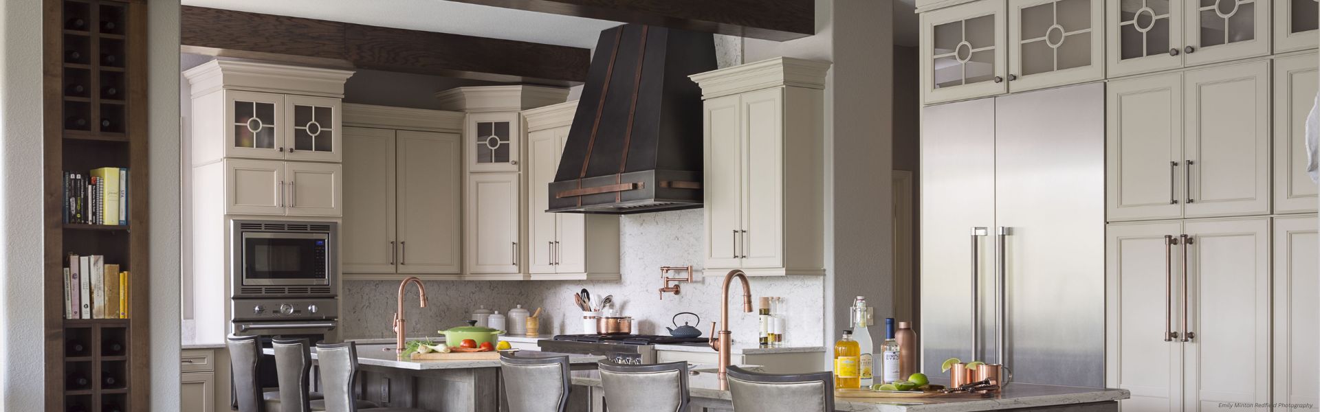 Modern kitchen featuring Banner Vail Custom light beige cabinets, stainless steel appliances, a large refrigerator, black range hood, island seating with gray chairs, counters with fruit and drinks, and shelves with books on the left.