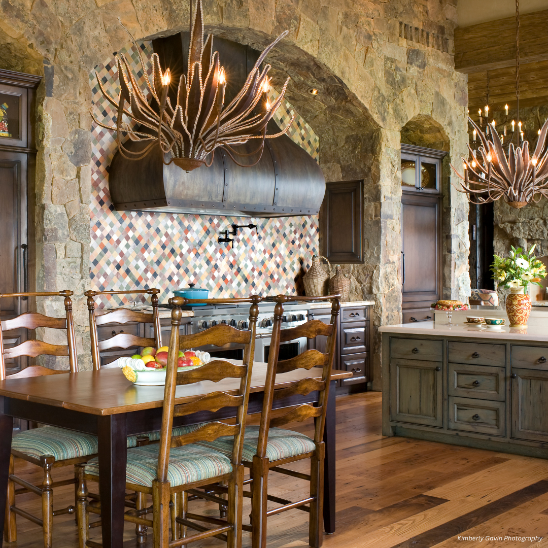 Bellvue gallery2 The Bellvue gallery2 features a rustic kitchen with stone walls, wooden cabinets, tiled backsplash, wood dining table and chairs, island adorned with flowers, and antler-shaped chandeliers for a natural yet elegant look.