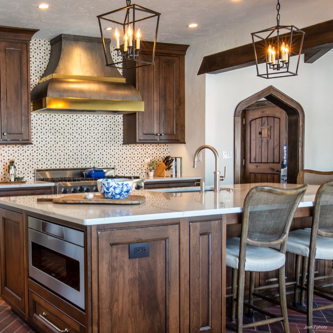 The Bonner Peak gallery1✅ features a modern kitchen with wooden cabinets, a spacious island with bar stools, stainless steel range hood, patterned backsplash, pendant lights, and a distinctive arched wooden door in the background.