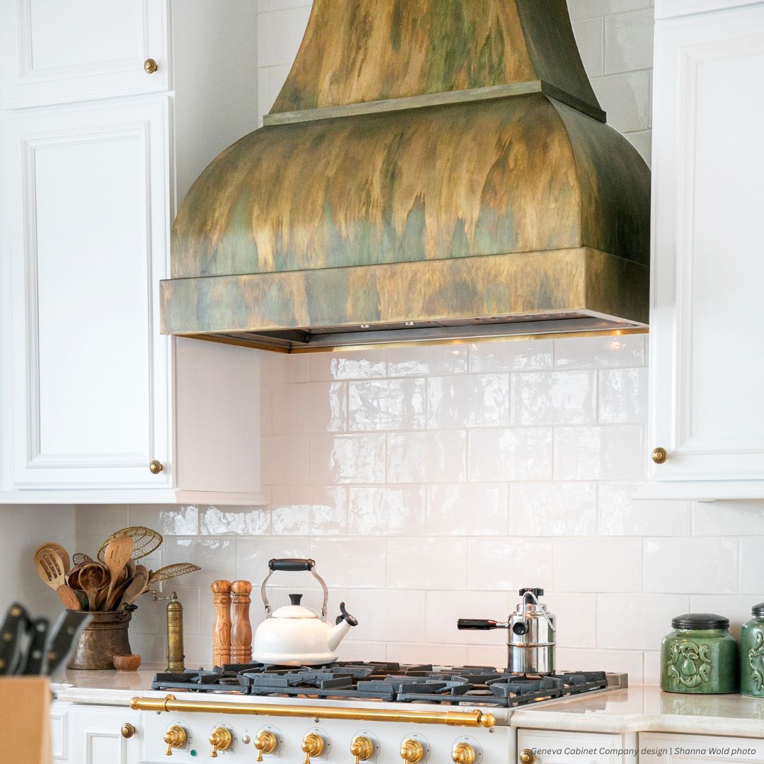The Bonner Peak gallery3 stove with gold knobs, a white kettle, and a coffee maker sits below a patina-finished range hood. White cabinets and glossy subway tile backsplash complete this bright, modern kitchen.
