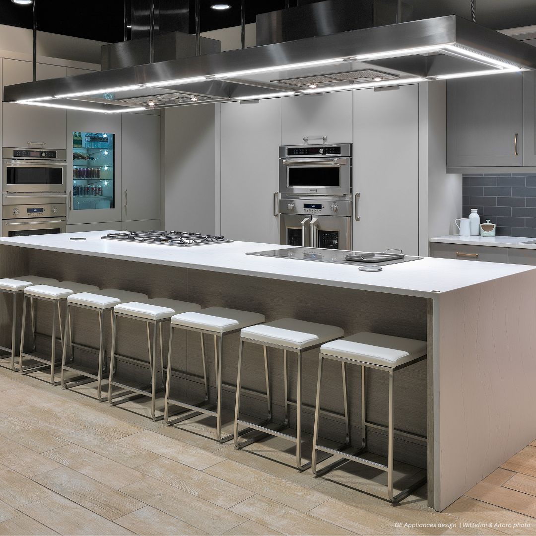 The Boulder gallery1✅ features a modern kitchen with a large island, six white cushioned bar stools, stainless steel appliances, built-in ovens, gas cooktop, and sleek gray cabinetry with under-cabinet lighting.