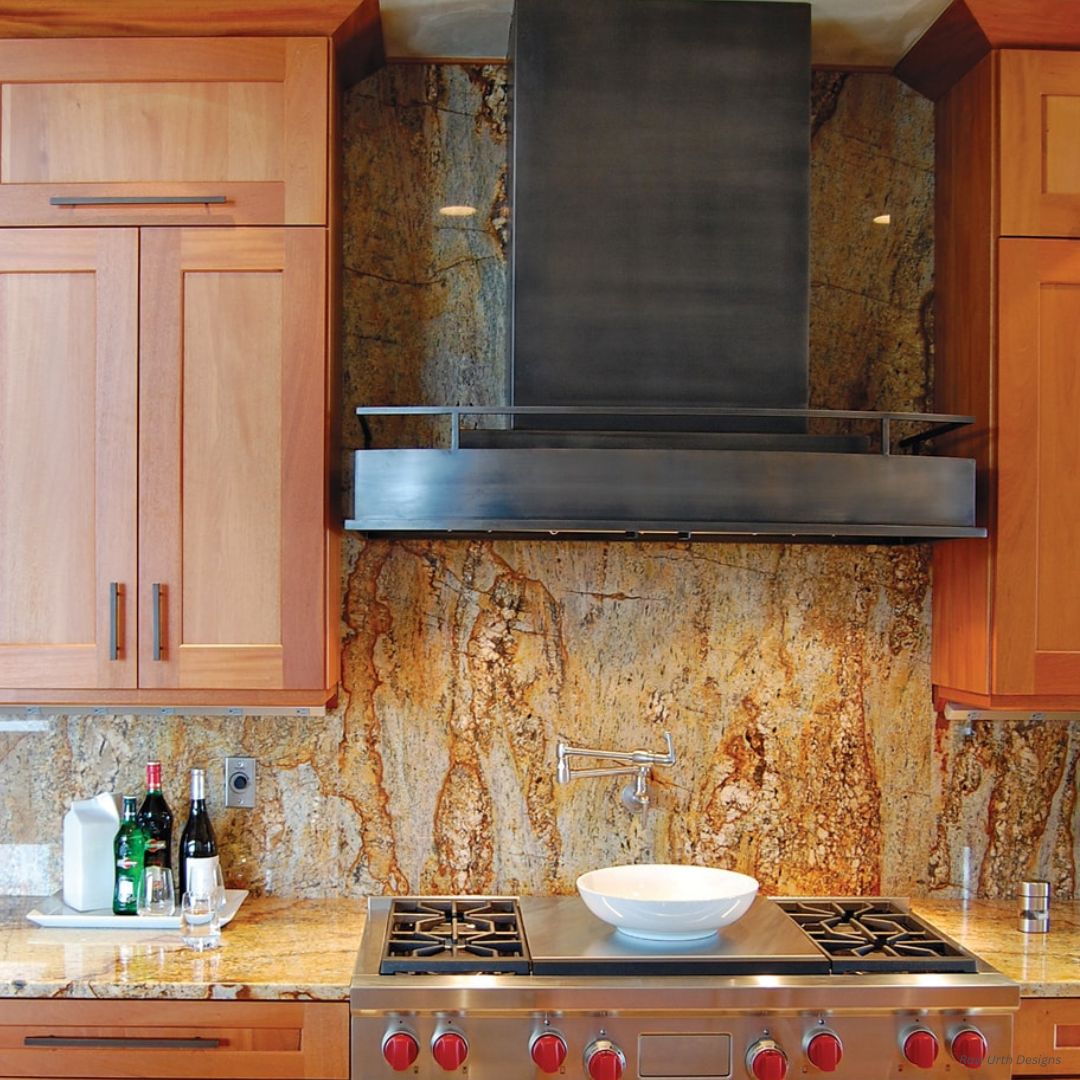 The Boulder gallery3✅ features a modern kitchen with a natural stone backsplash, wooden cabinets, a stainless steel stovetop with red knobs, black range hood, and a countertop displaying wine bottles, glasses, and a paper towel holder.