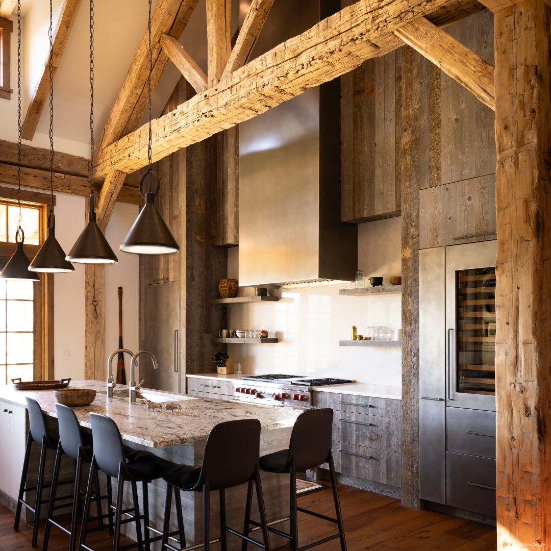 Box gallery2✅ Box gallery2✅ features a rustic kitchen with exposed wooden beams, a stone island with sink and four black barstools, industrial pendant lights, stainless steel appliances, gray wood cabinetry, and warm natural light throughout.