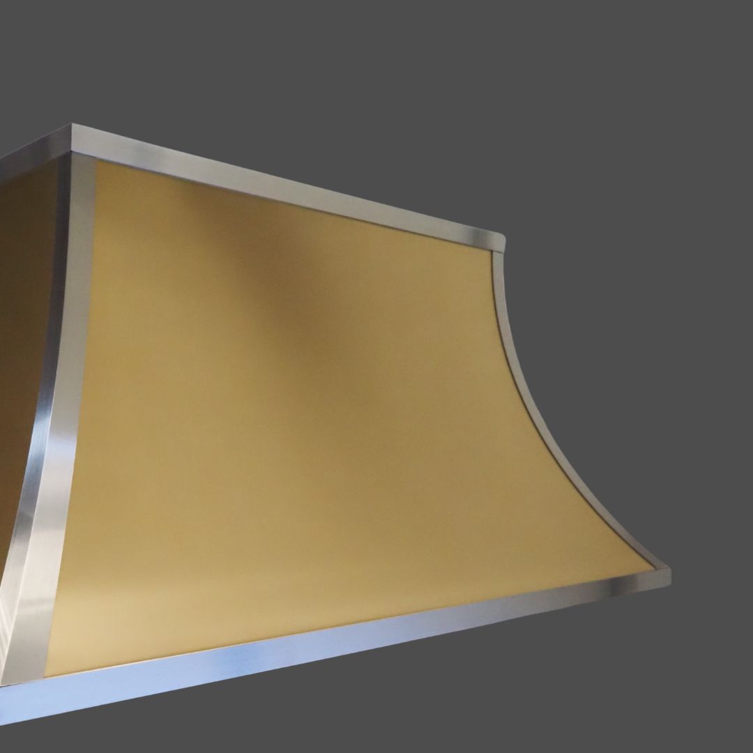 Brass Burnished 3 A close-up of the Brass Burnished 3 kitchen range hood features a gold-toned, curved surface with silver metallic trim, set against a plain gray background.