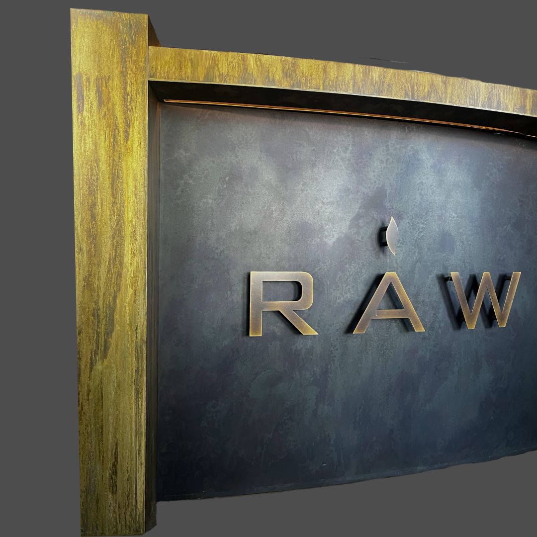 Brass Washed 2 A close-up of the Brass Washed 2 sign shows the partial word "RAW" in metallic letters on a dark textured background, framed by wood with a golden finish.